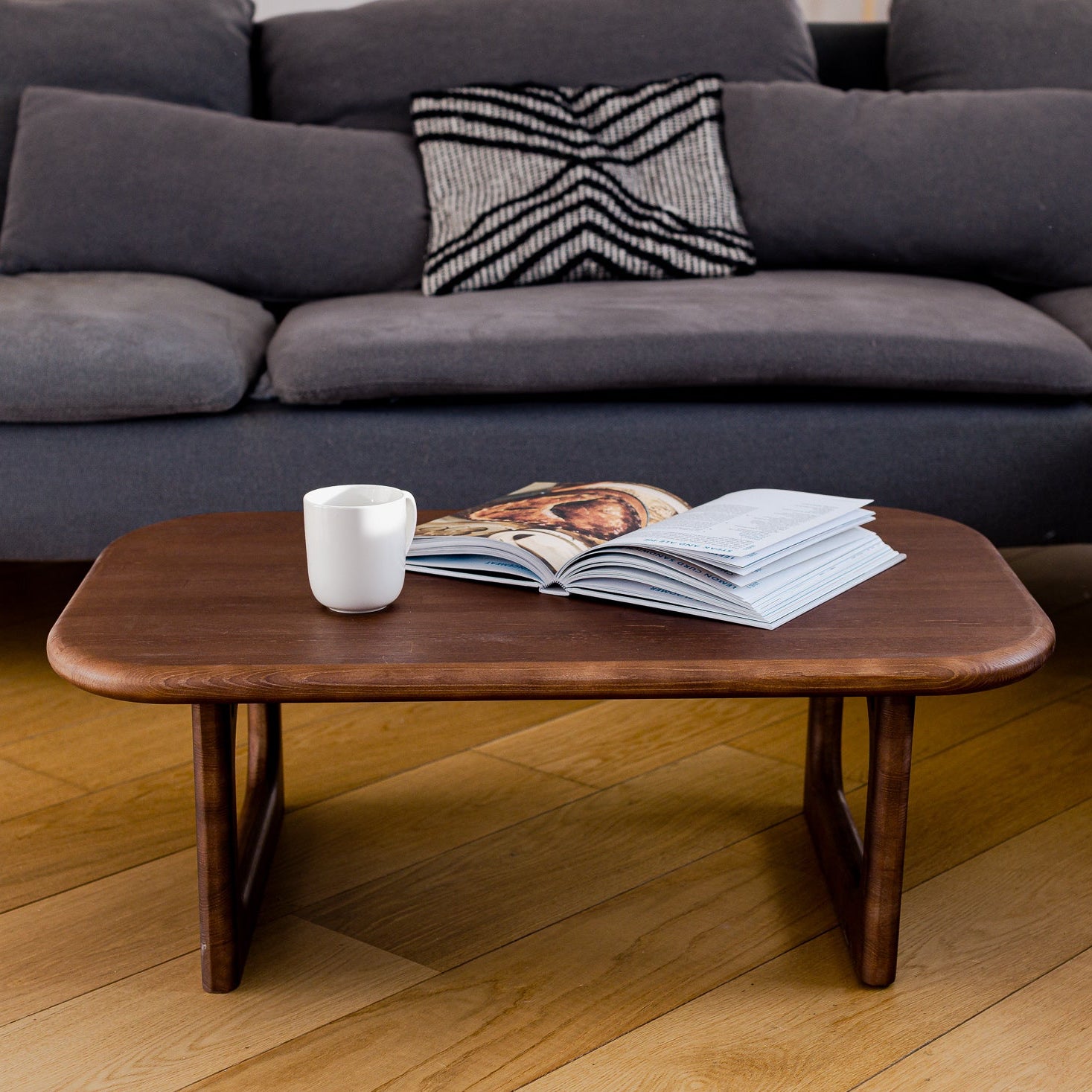 Gray sectional sofa with a wooden coffee table featuring a magazine and cup on a wooden floor.